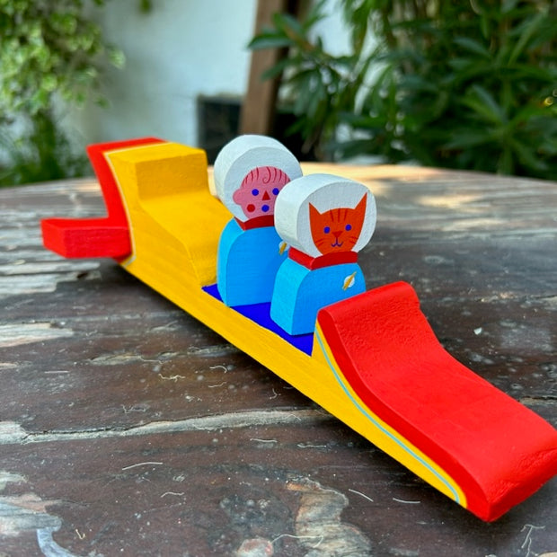 Brightly painted wooden sculpture of a long yellow and red space shuttle, with an open air seating. A person wearing a space suit and a helmet and a cat, dressed the same, sit in the cockpit.