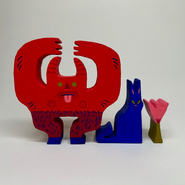 Wooden die cut sculptures of a red devil holding a flower over its head and a blue cat, sitting nearby. The devil has horns, green eyes, its tongue out and hairy goat legs.