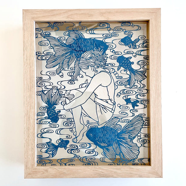 Intricate paper cutting on indigo colored paper of a girl, curled up with her knees drawn in, laying down. Around her are many fish swimming and a swirled water pattern. Piece is in a wooden frame.