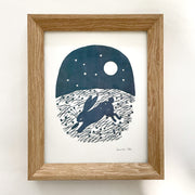 Simplistic outdoor night scene made of cut paper of a rabbit running with a carrot in its mouth. A full moon and stars are overhead and long stars and grass are on the ground. Paper is navy blue on a white background and in a wooden frame.