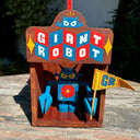 Painted wooden showcase box that holds a blue wooden robot inside. Exterior reads "Giant Robot" written in stylistic retro font, red blue and white. Wooden robot has a flower on its stomach and holds a yellow flag.
