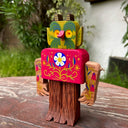 Wooden sculpture of a forest creature with tree trunk legs, a rounded square body and a green face with yellow eyes, leaf pattern ears and small horns. Its face, body and arms have delicately painted floral patterns on them, akin to folk art.