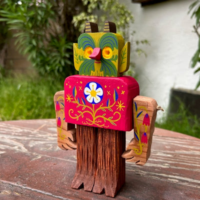 Wooden sculpture of a forest creature with tree trunk legs, a rounded square body and a green face with yellow eyes, leaf pattern ears and small horns. Its face, body and arms have delicately painted floral patterns on them, akin to folk art.
