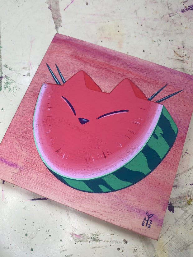 Painting of a slice of watermelon, with pointed ears like a cat and a closed eye cat expression. It is against a pink ombre wood grain background. Shown at an angle.