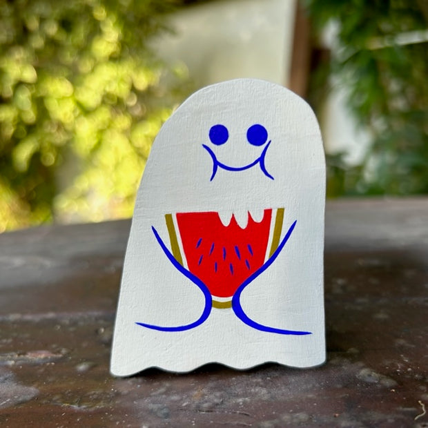 Die cut wooden sculpture of a white ghost, holding a partially bitten watermelon slice. Its mouth is closed and smiling with full cheeks.