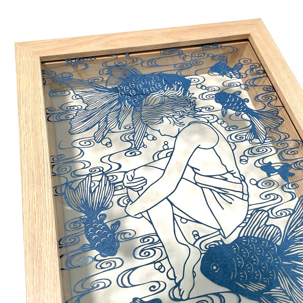 Intricate paper cutting on indigo colored paper of a girl, curled up with her knees drawn in, laying down. Around her are many fish swimming and a swirled water pattern. Side angle.