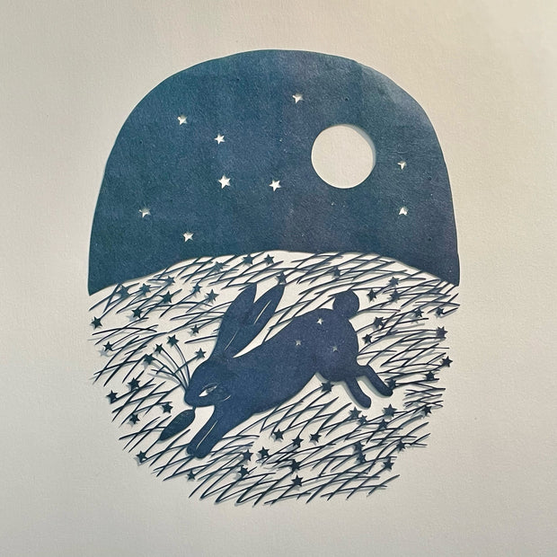 Simplistic outdoor night scene made of cut paper of a rabbit running with a carrot in its mouth. A full moon and stars are overhead and long stars and grass are on the ground.