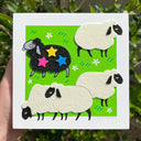 Illustration of felt sheep on a green, grassy background. 4 of the sheep are white and the other is black with 3 stars on it.