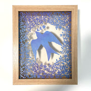 Purplish blue hand cut paper cut design of a bird carrying a single stemmed flower with a star shaped blossom. Around the bird is a pattern of leaves and flowers. Inside a transparent backed wooden frame.