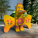 Painted assembled wooden sculpture of a yellow dinosaur like monster with wings, Rodan. It has a large beak with sharp teeth bared and pink and blue folk art style flowers on its wings and stomach.
