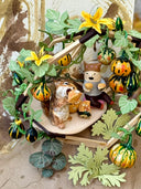 Diorama artwork of a chipmunk eating a gourd with a person, as they sit in a shelter made out of wood sticks and with gourd vines wrapped all around.