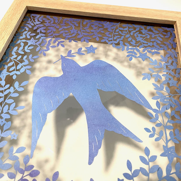 Purplish blue hand cut paper cut design of a bird carrying a single stemmed flower with a star shaped blossom. Around the bird is a pattern of leaves and flowers. Inside a transparent backed wooden frame.