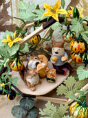 Diorama artwork of a chipmunk eating a gourd with a person, as they sit in a shelter made out of wood sticks and with gourd vines wrapped all around.