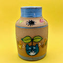 Stoneware vase featuring small, cute illustrations of chibi blue Totoros, holding leaf sprouts with acorns, mushrooms and leaves nearby. Small dust sprites go around the top of the blue lined vase.