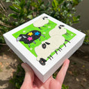 Illustration of felt sheep on a green, grassy background. 4 of the sheep are white and the other is black with 3 stars on it.