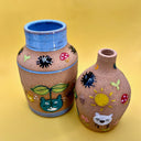 Stoneware vase featuring small, cute illustrations of chibi blue Totoros, holding leaf sprouts with acorns, mushrooms and leaves nearby. Small dust sprites go around the top of the blue lined vase.