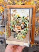 Diorama artwork of a chipmunk eating a gourd with a person, as they sit in a shelter made out of wood sticks and with gourd vines wrapped all around.