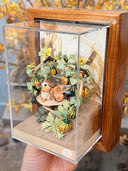 Diorama artwork of a chipmunk eating a gourd with a person, as they sit in a shelter made out of wood sticks and with gourd vines wrapped all around.