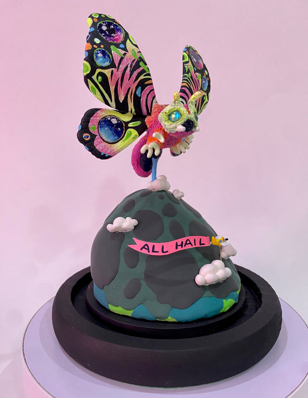 Mixed media sculpture of a large psychedelically colored moth, with large eyes hovering over a rounded land mass. A small helicopter flies around the mound with a banner that reads "All Hail"