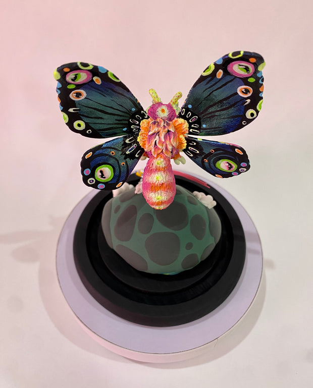 Mixed media sculpture of a large psychedelically colored moth, with large eyes hovering over a rounded land mass. A small helicopter flies around the mound with a banner that reads "All Hail"