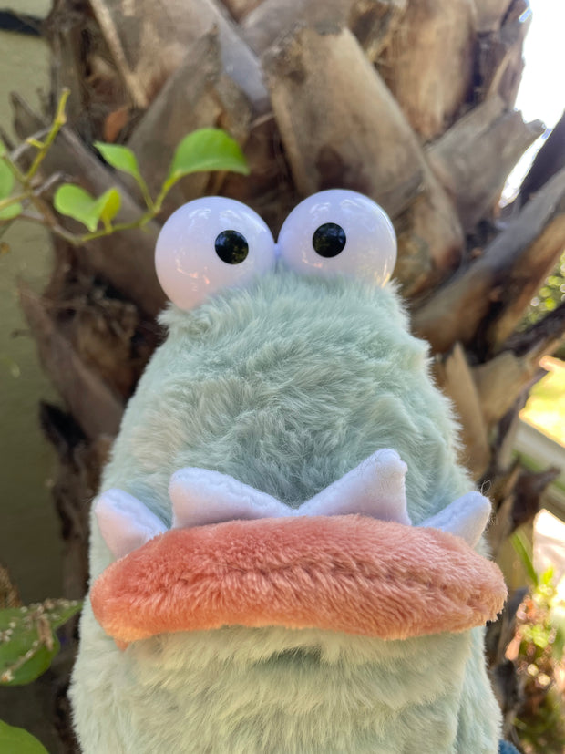 Light green plush of a goofy looking monster creature, with 2 eyes atop its head and a big underbite. It has 3 fins, 2 on each side of its body and one on the back of its tail, like a seal.