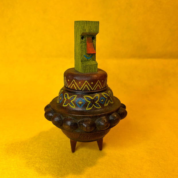 Decorative wooden object, shaped loosely like UFO with a colorful green Tiki head figure