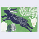 Green rectangular fabric placemat with an illustration of a navy blue jackalope, leaping in the air. Behind it is a stylistic white tree and under it are stylized rocks.