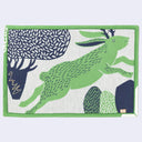 Green rectangular fabric placemat with an illustration of a navy blue jackalope, leaping in the air. Behind it is a stylistic white tree and under it are stylized rocks. Back view shows inverted colors.