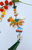 Decorative hanging with a brown horse clay sculpture covered in fruit stickers. Hanging also includes smaller charms: a tangerine, cup of coffee, car and dog with a green tassel on a light blue background