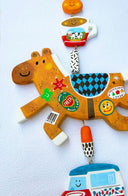 Decorative hanging with a brown horse clay sculpture covered in fruit stickers. Hanging also includes smaller charms: a tangerine, cup of coffee, car and dog with a green tassel on a light blue background