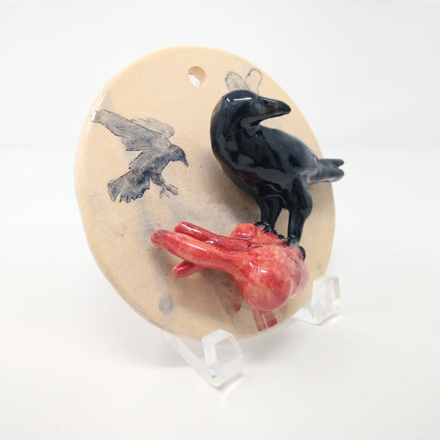 Ceramic sculpture of a cream colored circle disk, with a black crow standing atop of a bleeding human heart. Both the crow and the heart are 3D and jut out from the circle. Shown at the side to display three dimensionality. 