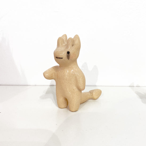 An off-white ceramic sculpture of a small 4 horned devil, with subtle tears. It has a hand extended out and a thick devil tail.