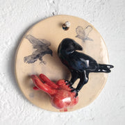 Ceramic sculpture of a cream colored circle disk, with a black crow standing atop of a bleeding human heart. Both the crow and the heart are 3D and jut out from the circle.