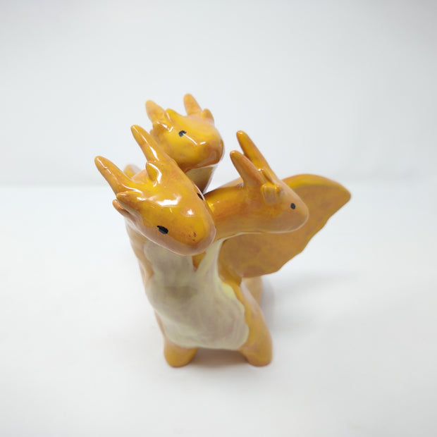 Ceramic sculpture of King Ghidorah, a 3 headed orange dragon monster. It has simplistic body shapes and horns atop its heads. They crane in different directions.