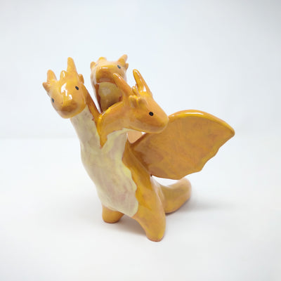 Ceramic sculpture of King Ghidorah, a 3 headed orange dragon monster. It has simplistic body shapes and horns atop its heads. They crane in different directions.
