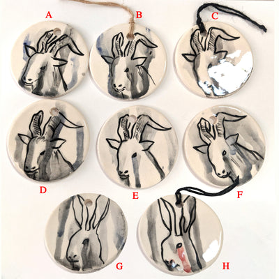 8 flat circular ceramic ornaments, 6 of have black line art paintings of horned goats with watercolor style drips. Bottom 2 feature black line art paintings of rabbits, with streaking watercolor tears.