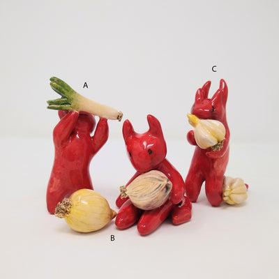 3 red ceramic devil characters with simplistic figures and detailing. One holds up a leek, another holds a cut onion with an onion sitting at its feet. The final one holds a bulb of garlic with another garlic bulb at its feet.
