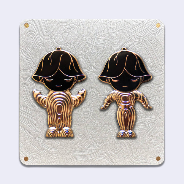 2 die cut gold outlined enamel pins on an elaborate cream color backing card. Both pins are of characters with upside down flowers atop their heads and simple closed eye smiles. Their bodies are abstract in shape and striped gold, with black heads.. 