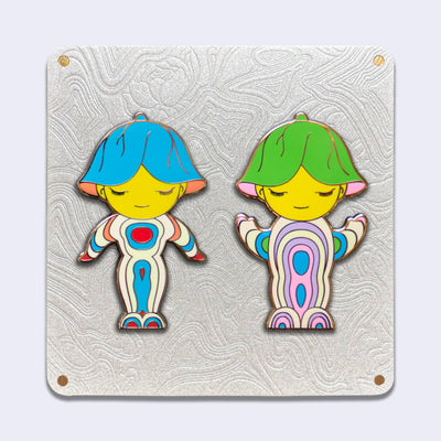 2 die cut gold outlined enamel pins on an elaborate cream color backing card. Both pins are of characters with upside down flowers atop their heads and simple closed eye smiles. Their bodies are abstract in shape and are strips of colors. Right one is blue, cream, orange and red. Right one is green, blue, cream, purple and pink.