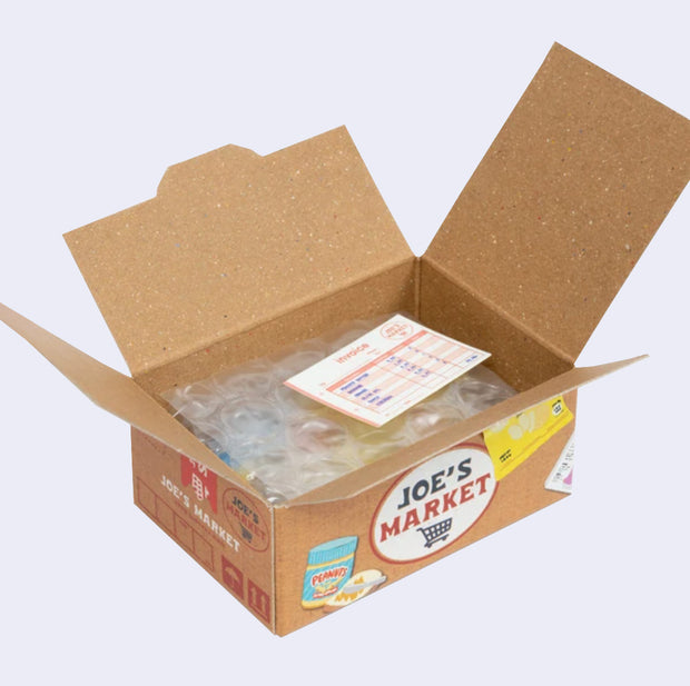 Open box of miniature stickers, made to resemble a shipping box with bubble wrap around the product and a small invoice. Exterior of box looks like realistic Trader Joe's imitation grocery box.