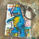 Blue and yellow dinosaur keychain with a crown on a clear plastic card.