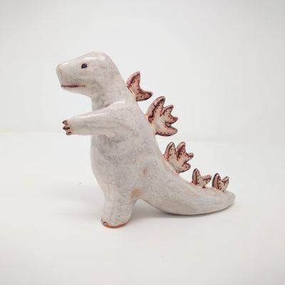 White ceramic sculpture of Godzilla, made of mostly smooth features with a simple line mouth and eyes drawn on. It has spikes running down its back and has one hand extended out. A rust brown is used as accent coloring.