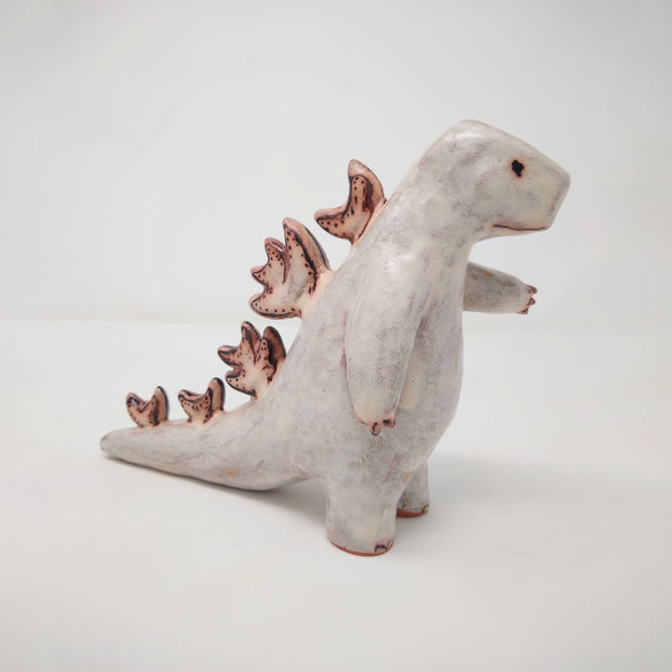 White ceramic sculpture of Godzilla, made of mostly smooth features with a simple line mouth and eyes drawn on. It has patterned spikes running down its back and has one hand extended out. A rust brown is used as accent coloring.