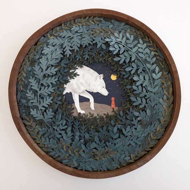 Layered cut paper diorama style sculpture in an open wooden circle frame. A white wolf looks down on a small person wearing a red jacket. Many leaves frame the scene.