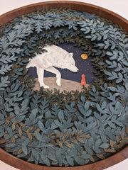 Layered cut paper diorama style sculpture in an open wooden circle frame. A white wolf looks down on a small person wearing a red jacket. Many leaves frame the scene.