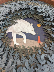 Layered cut paper diorama style sculpture in an open wooden circle frame. A white wolf looks down on a small person wearing a red jacket. Many leaves frame the scene.