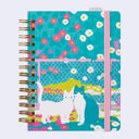 A spiral-bound notebook with a colorful cover featuring two cats in front of a fence, set against a turquoise background with pink and white flowers. It has a pink elastic closure band and gold-tone spiral binding.