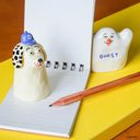 Two ceramic figures, one resembling a dog and the other a ghost, on a notebook with a pencil.