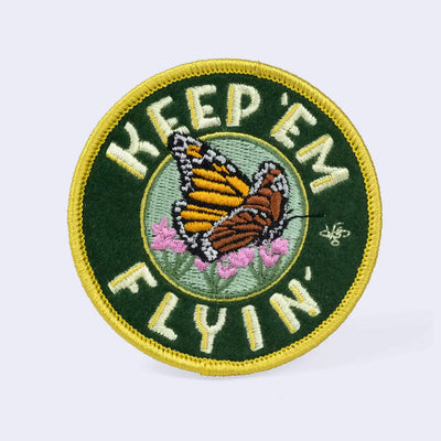 Dark green circular embroidered felt patch with a yellow border and retro font that reads "Keep Em Flyin" with an illustration of a monarch butterfly resting on pink flowers.