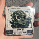 Hand holding a sticker with a stylized green creature illustration.